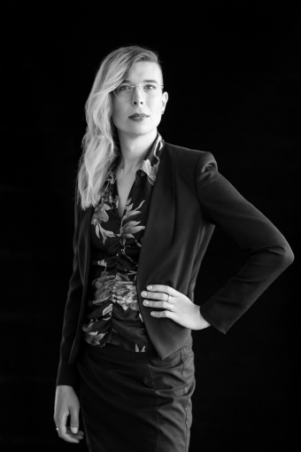 Grayscale photography of Janis Joan König.
            Janis is standing, with her left arm positioned on her waist, slightly tilted towards the viewer.
            She wears a dark blazer above a flowery fitted top and dark pencil skirt. Her hair is long and curly,
            falling to the right in front of her shoulder around her glasses, leaving her sidecut on the left exposed.